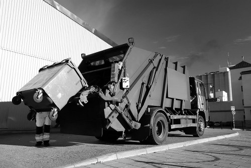 A large industrial rubbish collection vehicle, with a hydraulic lift arm extended, is positioned on a paved street outside a commercial building, ready for waste removal. The vehicle features a dark, metallic body with a smooth, reflective finish, and has multiple compartments for waste storage. In the foreground, a worker wearing high-visibility clothing and protective footwear is seen handling a large wheeled waste bin, which is being lifted towards the vehicle's hopper for disposal. The waste bin has a sturdy plastic body with a textured surface in a light tone, and is positioned at an angle as it is being emptied. The background includes a modern warehouse or industrial premises with corrugated metal walls and some distant factory structures. The scene is captured in natural daylight, adding clarity to the textures and colors of the equipment and environment. This setup illustrates typical commercial waste collection procedures performed by companies such as Commercial Waste Removal Chessington, serving local businesses in the Chessington area, within the postal district KX.