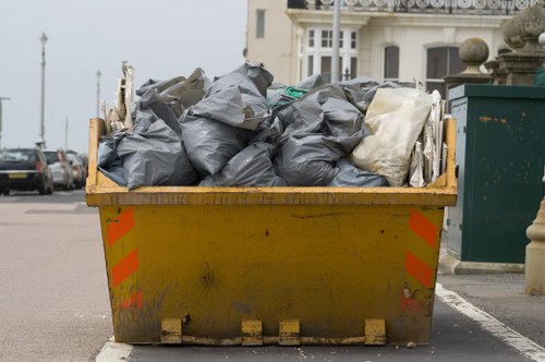 A large, industrial yellow skip bin filled with tightly packed black and white rubbish bags, situated on a paved street near residential buildings in Chessington. The bags appear to be made of durable plastic with a glossy finish, some wrinkled and creased, indicating recent disposal. The skip is positioned close to a curb, with the surrounding environment including parked cars, a green waste collection bin, and a building with large windows and decorative stone elements visible in the background. The scene is captured during daylight, with overcast weather creating even lighting, emphasizing the textures of the rubbish bags and the weathered surface of the skip. The image exemplifies typical commercial waste removal practices, with Commercial Waste Removal Chessington providing a clear visual of efficient rubbish clearance in a suburban setting, suitable for waste management services in the area.