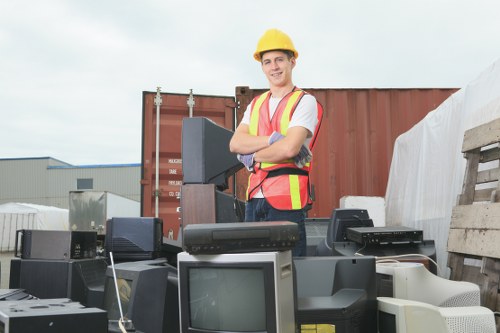 A young male worker wearing a yellow safety helmet and a high-visibility vest stands outdoors in an industrial environment with a clear sky overhead. He is smiling and has his arms crossed, positioned behind a collection of discarded electronic devices, including old computer monitors and towers, arranged on the ground. In the background, there is a large red shipping container, wooden pallets, and a section of a warehouse or industrial buildings, indicating a recycling or waste collection yard. The scene suggests a commercial rubbish removal context, with a focus on electronic waste disposal. The environment appears organized and typical of a professional waste management operation near Chessington, with natural daylight illuminating the scene.