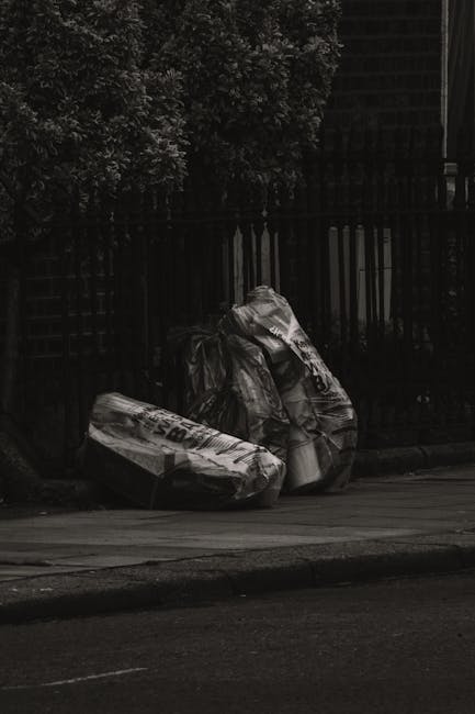 A large accumulation of black plastic rubbish bags filled with waste is stacked against a modern tiled building exterior. Some bags are tightly knotted, while others appear more loosely gathered. Seve