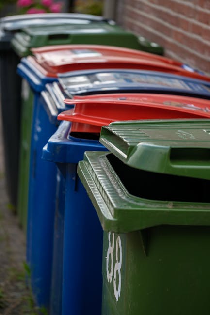 Close-up of a person wearing orange protective overalls and a white glove, holding a blue plastic rubbish bag tightly by its gathered top. The bag appears filled with waste and has a shiny, semi-trans
