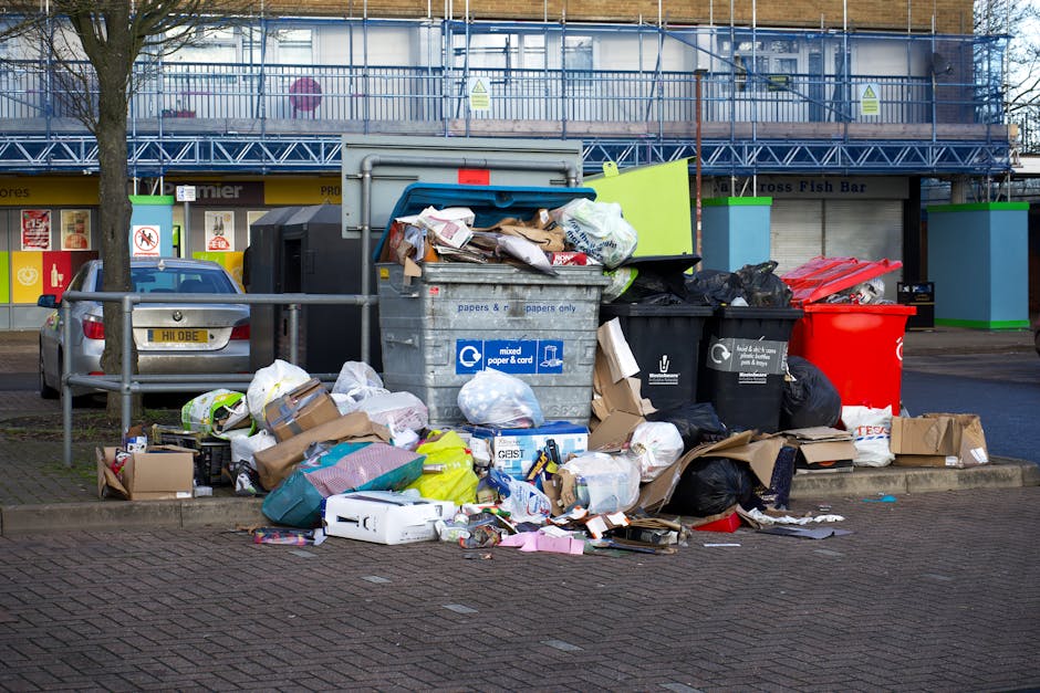 A small blue flatbed truck parked alongside a road, loaded with a variety of waste materials including cardboard boxes, plastic bags, and miscellaneous debris. The truck's sides are black with metal r